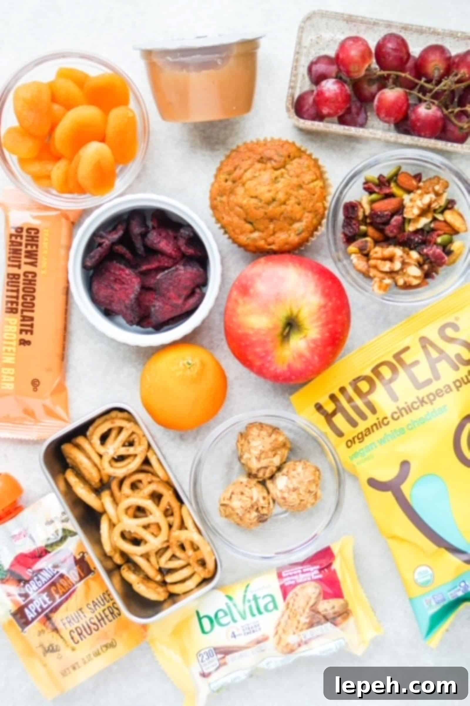 Individual snacks laid out on a surface: dried apricots, applesauce, grapes, beet chips, muffin, apple, trail mix, protein bar, spelt pretzels, HIPPEAS, energy bites, belVita package, and a apple-carrot fruit crusher from Trader Joe's.
