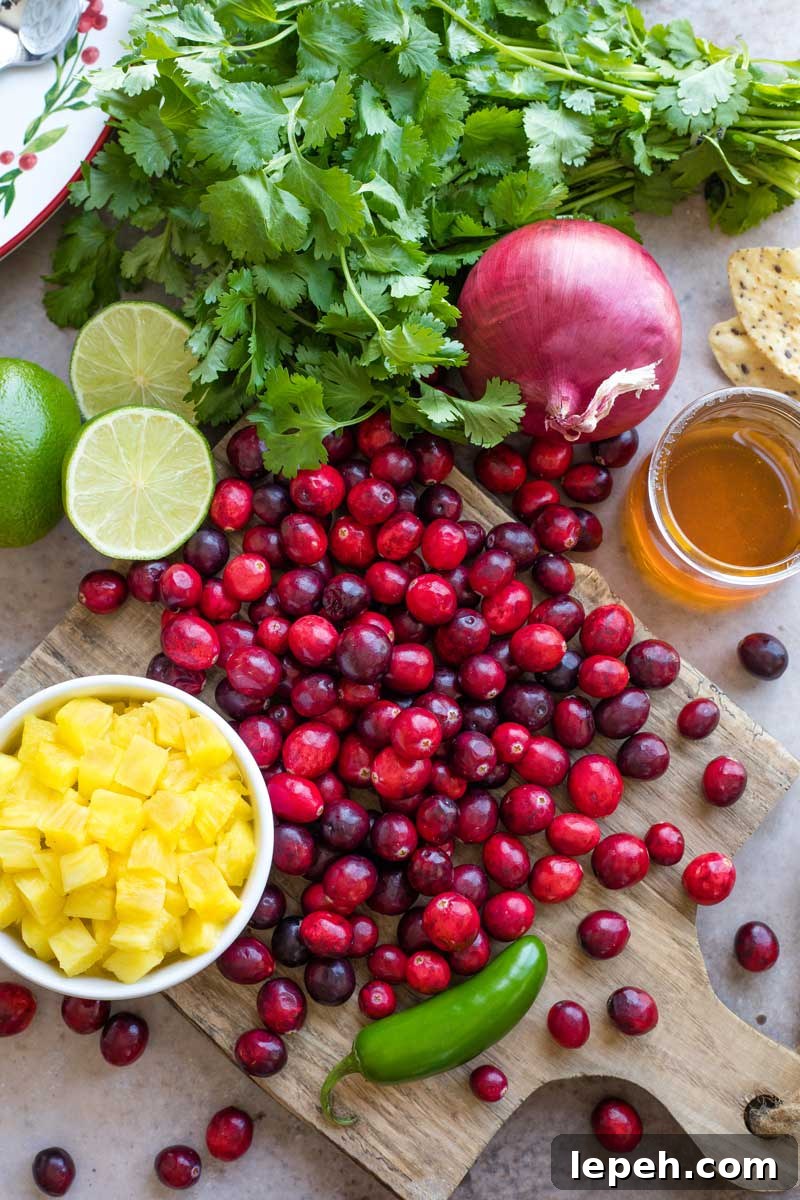 Overhead of ingredients for the salsa: a cutting board with a cascade of cranberry, a bowl of chopped pineapple, limes, jalapeno, honey and cilantro.,