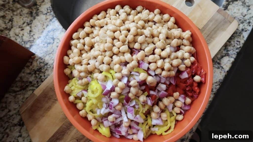 Adding the diced red onion, sliced banana peppers, roasted red peppers, diced provolone, diced charcuterie meat (such as salami, soppressata, and/or pepperoni), and chickpeas to the bowl with the pasta