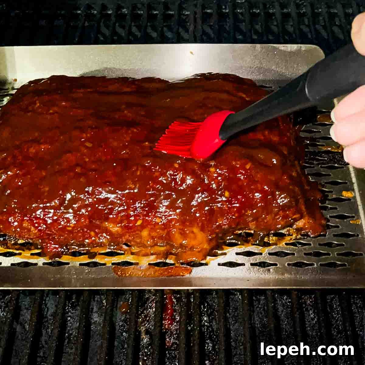 A red silicone basting brush adding barbecue sauce as a glaze to the smoked meatloaf.