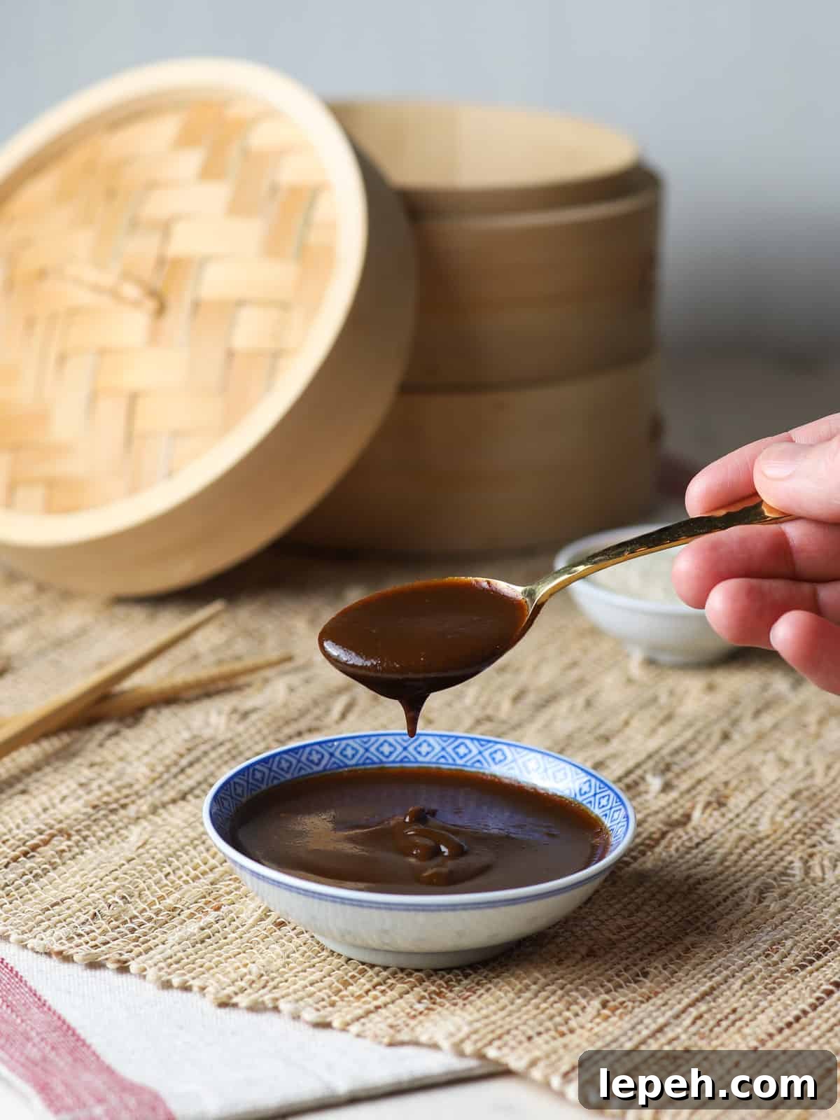 Photo of a shallow dish of hoisin sauce on a jute mat with someone holding a dripping spoon of sauce above the bowl and chopsticks and a bamboo steamer in the background.