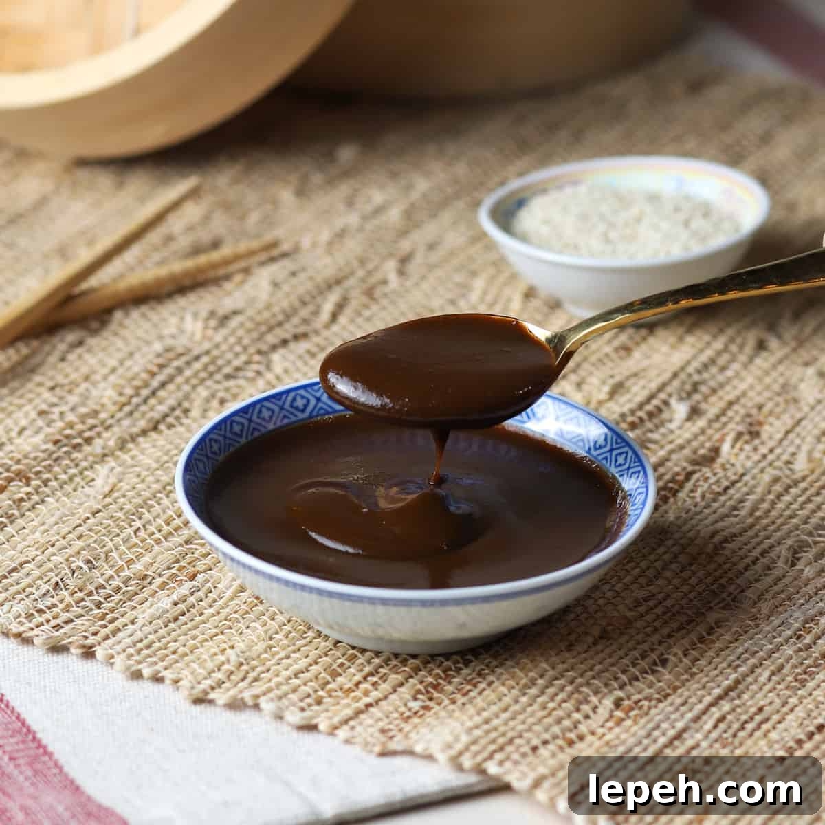 Photo of a Chinese shallow dish of hoisin sauce with a gold teaspoon held above dripping hoisin sauce.
