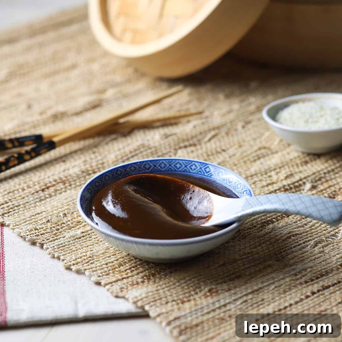Photo of a shallow Chinese bowl filled with homemade hoisin sauce and a Chinese ceramic spoon, all on a jute mat with bamboo steamer and chop sticks in the background.
