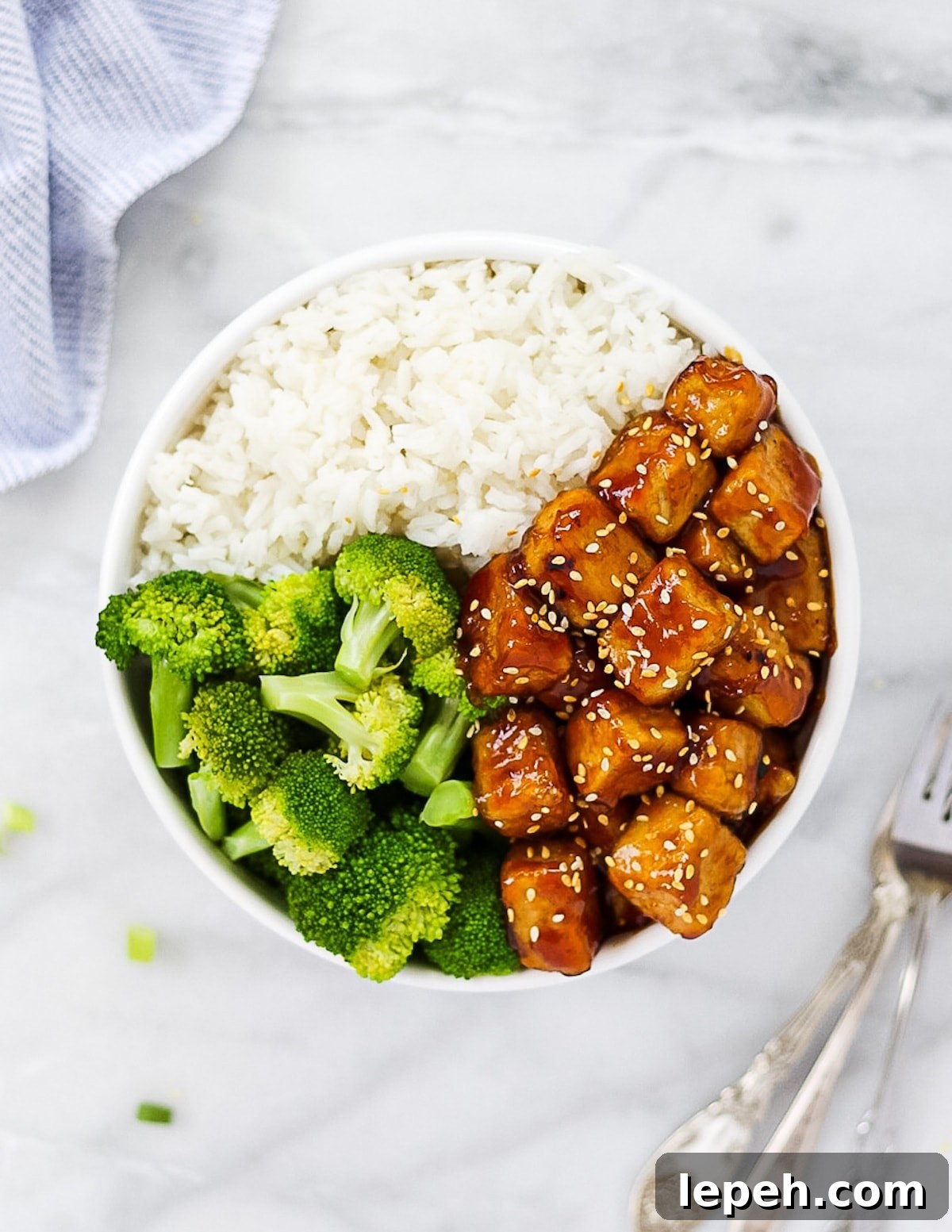 Overhead picture of glazed sesame tofu in a white bowl with rice and broccoli florets.