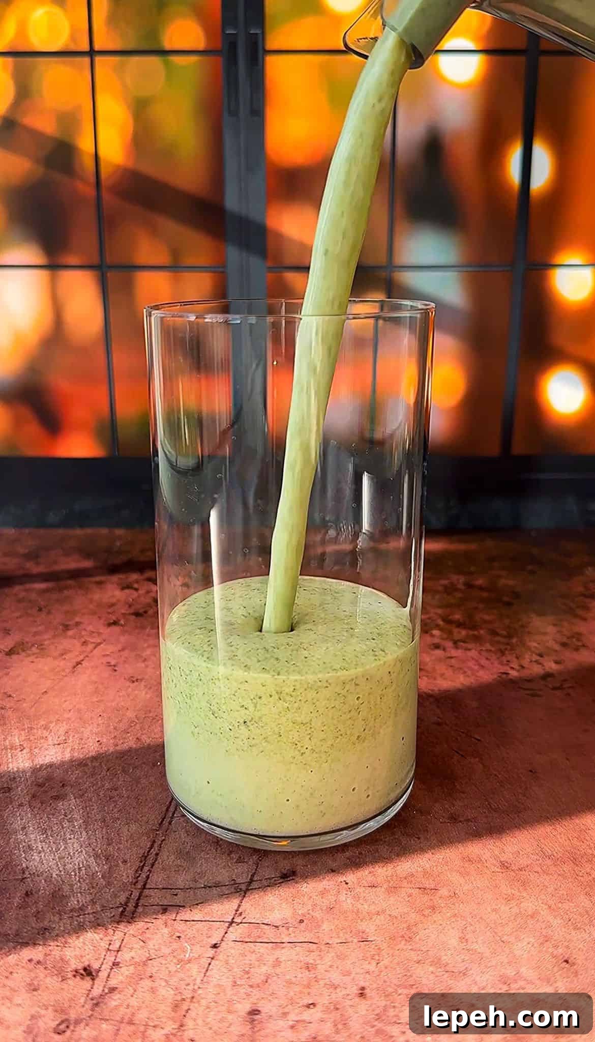 A thick, green smoothie is being poured into a clear glass. The background features a warm, out-of-focus display of glowing lights, creating a cozy ambiance. The scene is set on a wooden surface.