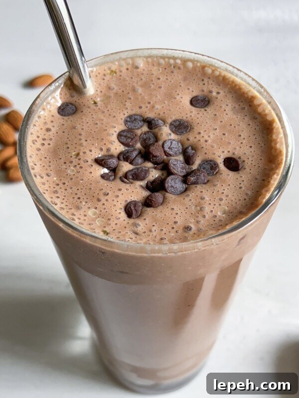 A glass of chocolate smoothie topped with chocolate chips, served with a metal straw. Some whole almonds are scattered in the background on a light surface.