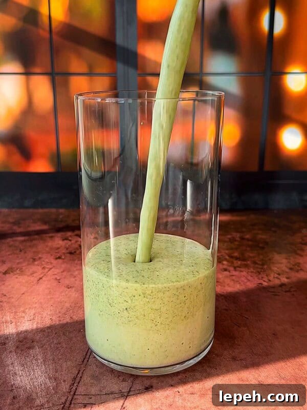 A thick, green smoothie is being poured into a clear glass. The background features a warm, out-of-focus display of glowing lights.