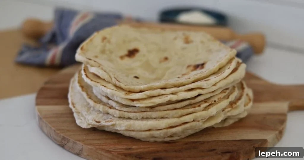 A stack of sourdough tortillas displayed on a round wooden serving board. You can see a wooden rolling pin and bowl of flour in the background.