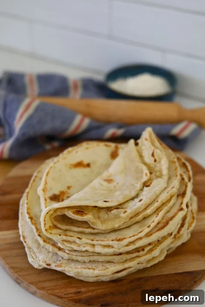 A stack of sourdough tortillas displayed on a round wooden serving board. You can see a wooden rolling pin and bowl of flour in the background.