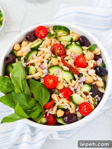 A white bowl sitting on top of a blue and white dish cloth. Inside the bowl is a Mediterranean Orzo Salad with orzo, chickpeas, tomato, olives, sun-dried tomatoes, red onion, and fresh herbs.