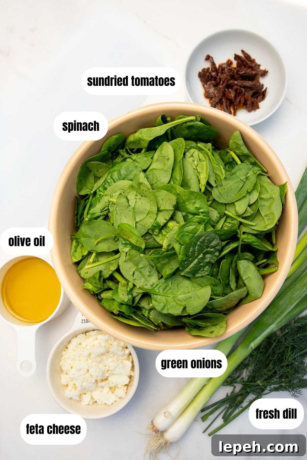 All ingredients to make Greek quesadillas on a white background including a bowl of fresh spinach, crumbled feta, green onions, fresh dill, olive oil and sundried tomatoes.