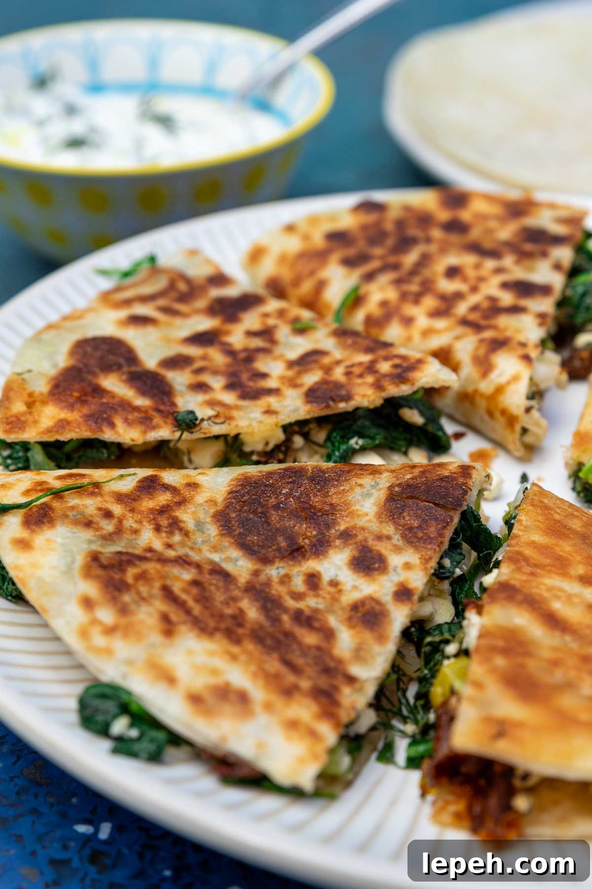 Closeup of Greek quesadilla triangles on a white plate and blue background with tzatziki sauce in the background.