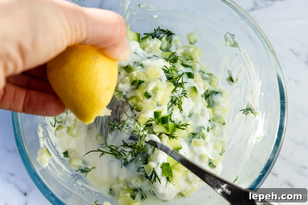 closeup of someone squeezing half a lemon into a bowl of all the ingredients for tzatziki in a glass bowl with a spoon from above.