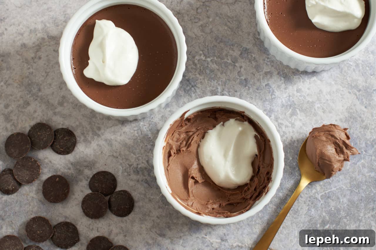 Three chocolate pots de creme with whipped cream on top. Chocolate wafers are on the left, a spoon with chocolate on it is on the right.