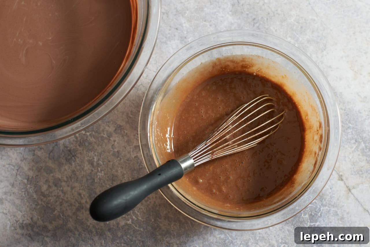 A whisk in a bowl of eggs mixed with warm chocolate. A large bowl of melted chocolate is on the left.