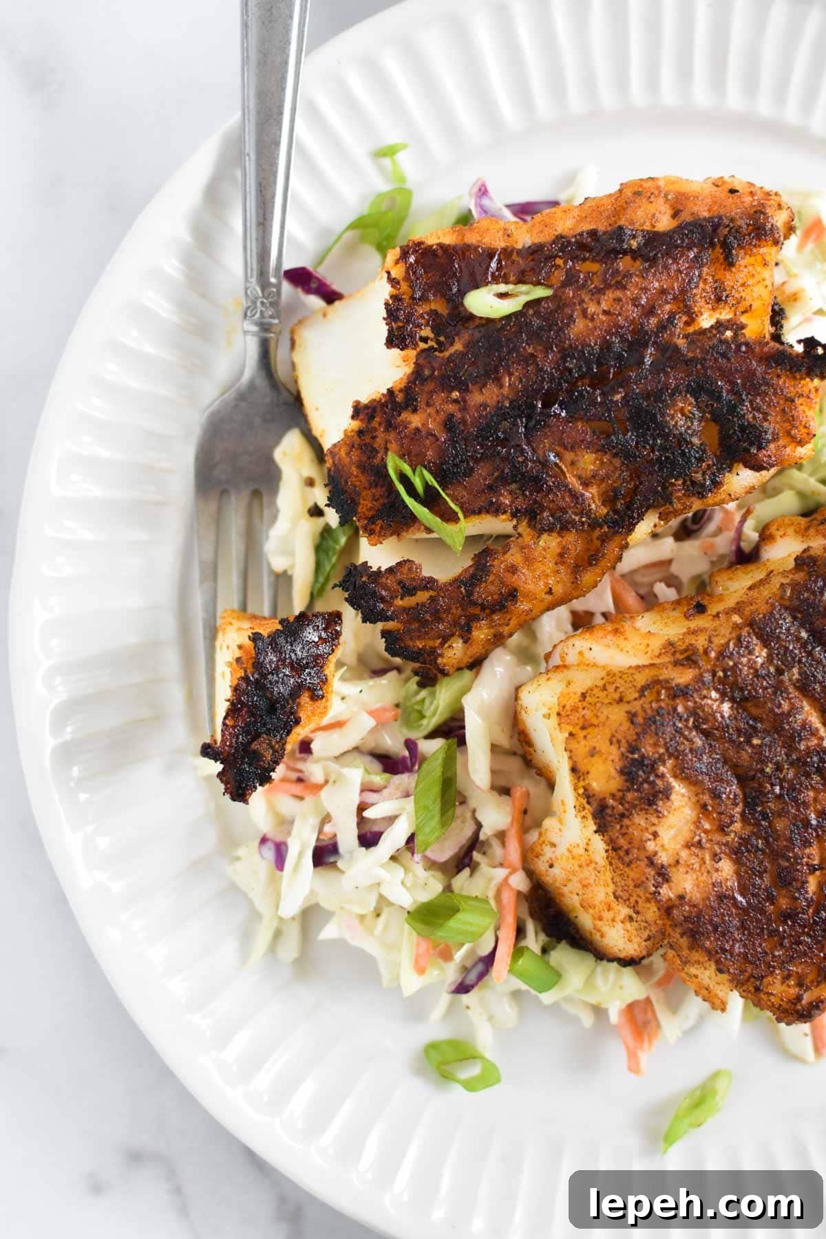 Two filets of blackened cod on top of a bed of coleslaw with a fork holding a piece of fish