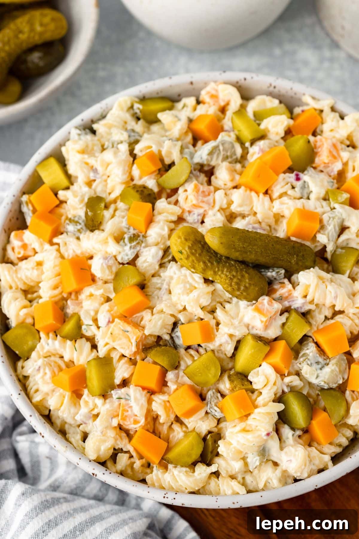 A bowl of creamy pasta salad with diced cheddar cheese, chopped pickles, and whole pickles on top, served on a light surface with extra pickles in the background.