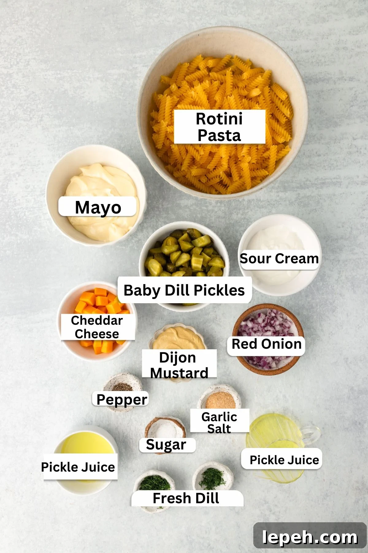 A top-down view of labeled ingredients for a pasta salad, including rotini pasta, mayo, sour cream, baby dill pickles, cheddar cheese, red onion, Dijon mustard, pepper, garlic salt, sugar, pickle juice, and fresh dill.