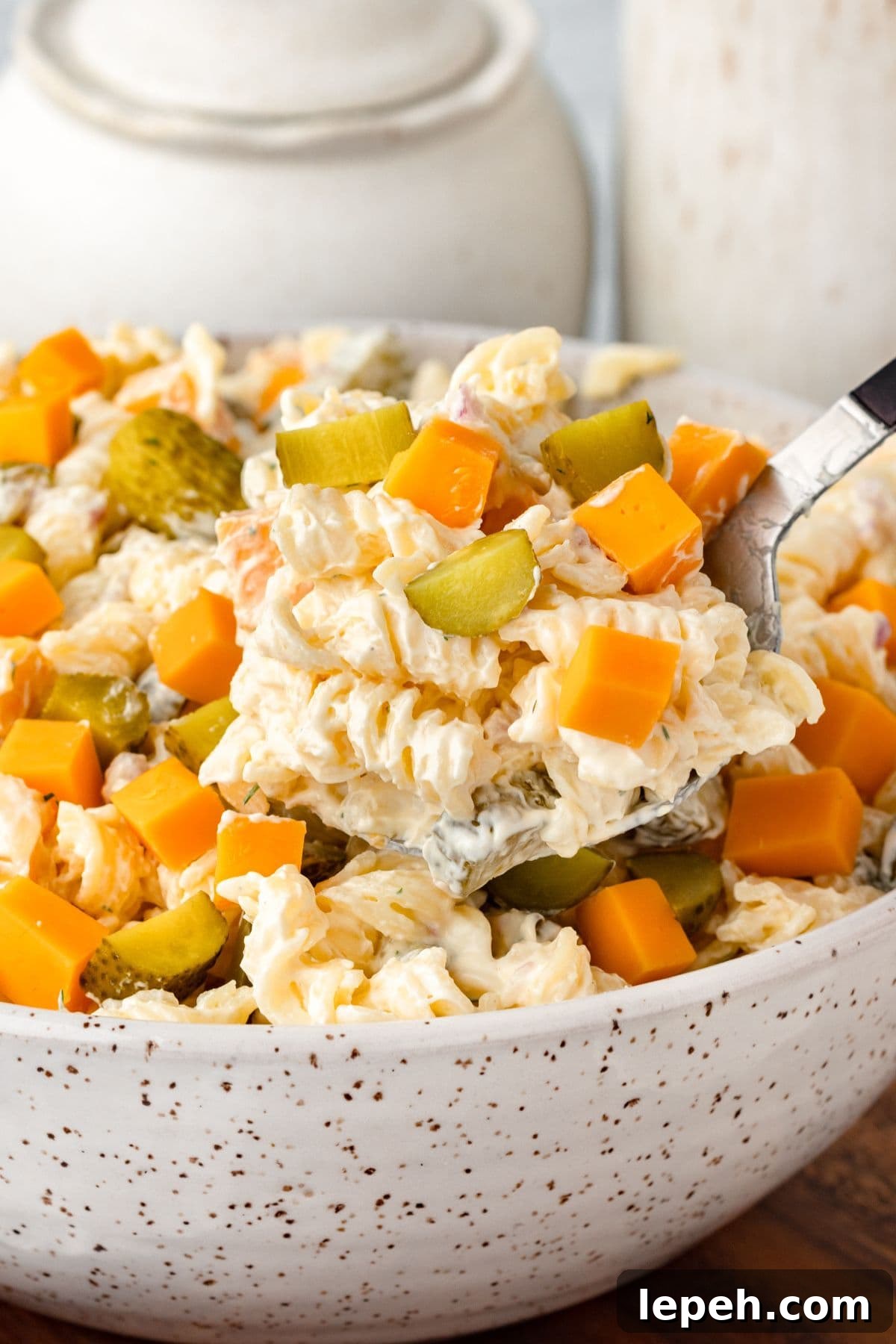 A close-up of a creamy pasta salad in a white speckled bowl, topped with cubed cheddar cheese and sliced pickles, with a serving spoon lifting a portion.