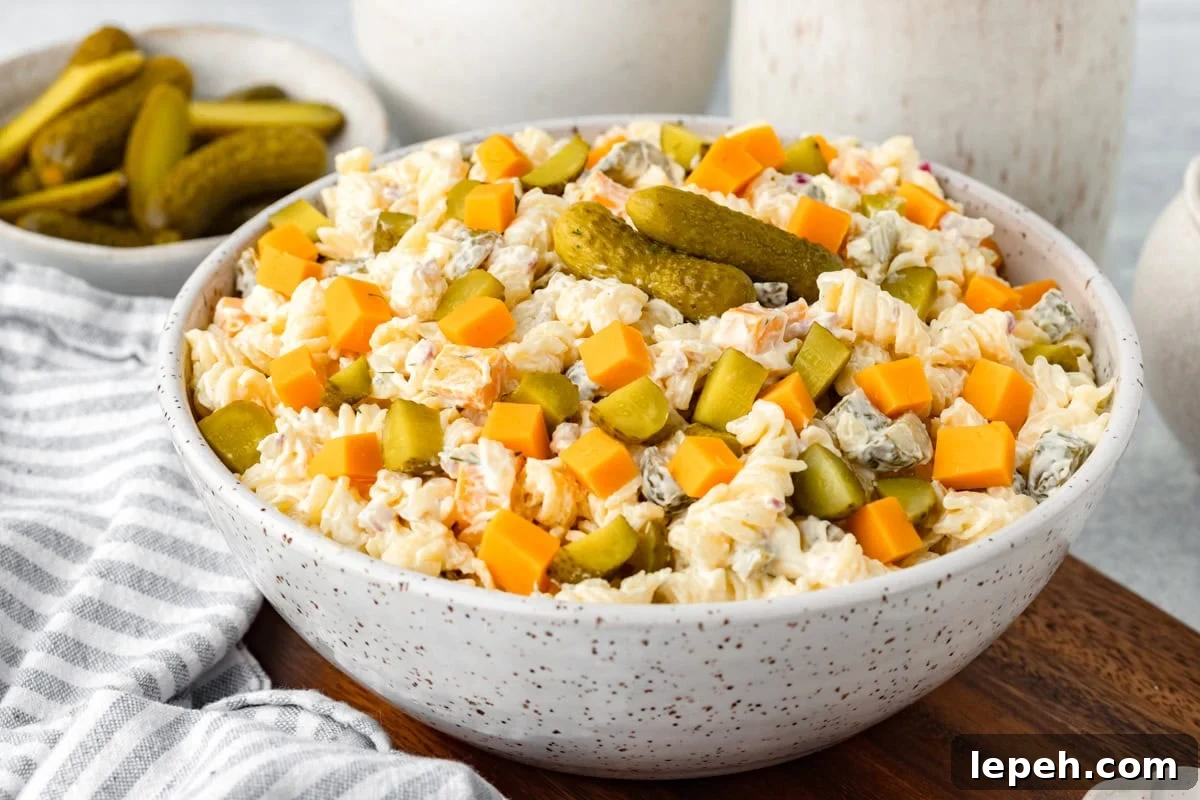A bowl of creamy pasta salad topped with diced cheddar cheese, sliced pickles, and whole gherkins, sits on a wooden board next to a striped napkin and a small bowl of extra pickles.
