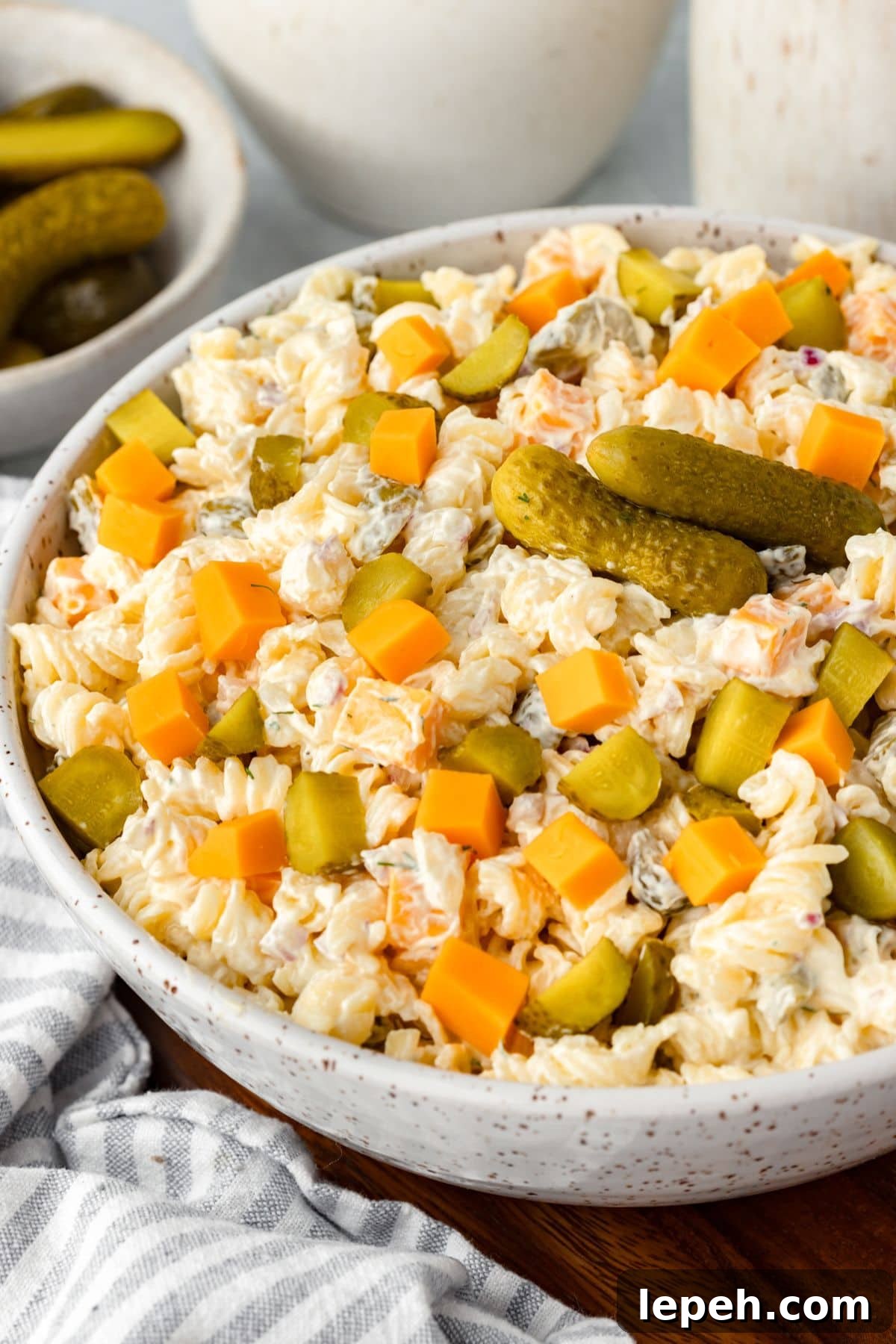 A bowl of creamy pasta salad with spiral noodles, cheddar cheese cubes, chopped pickles, and whole pickles on top, set on a striped cloth with a bowl of pickles in the background.
