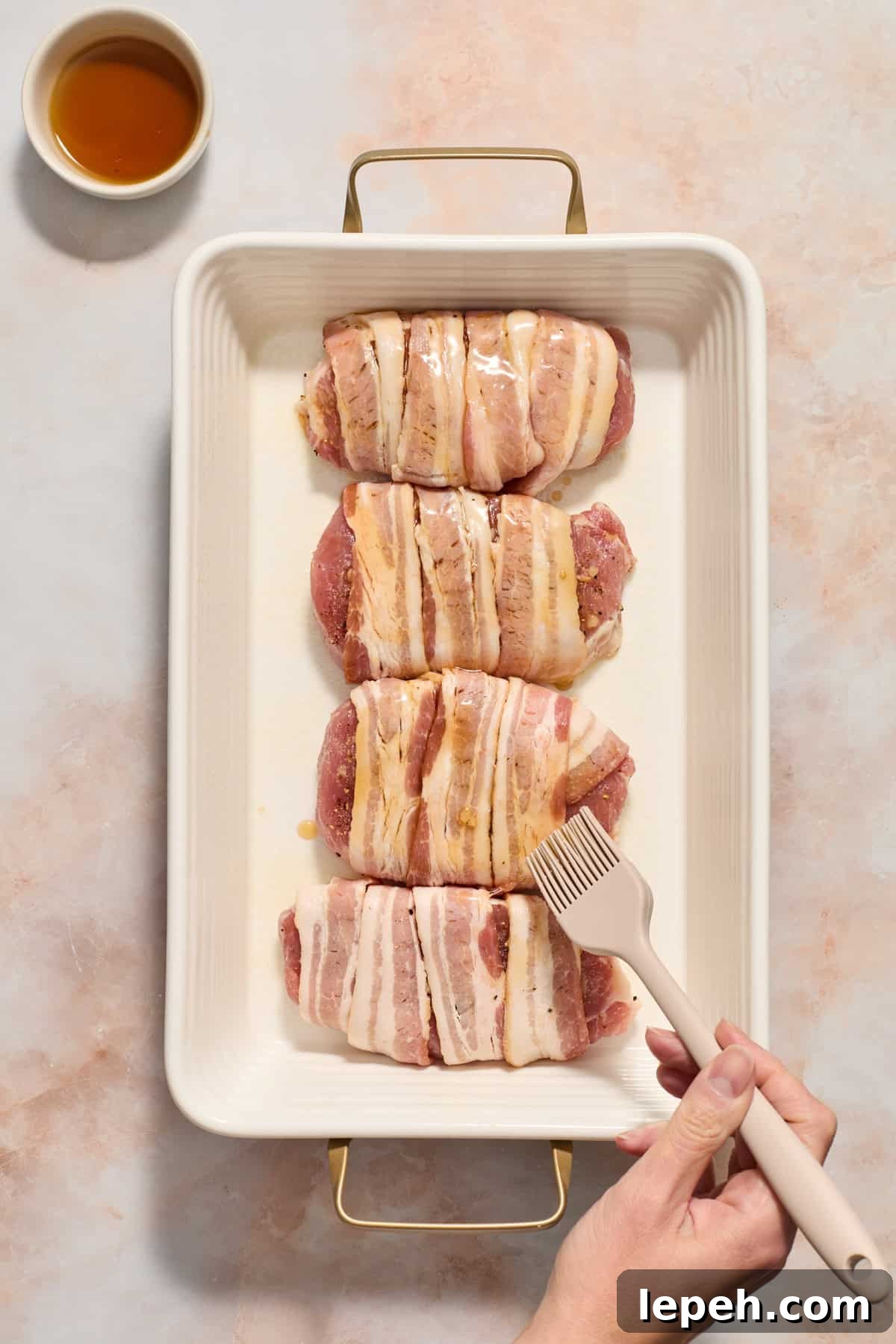 Maple being brushed on bacon wrapped pork chops in baking dish with dish of maple syrup beside the pan.